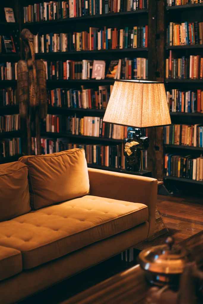 image features a dimly lit home library with mid century modern decor, yellow sofa, and lamp.